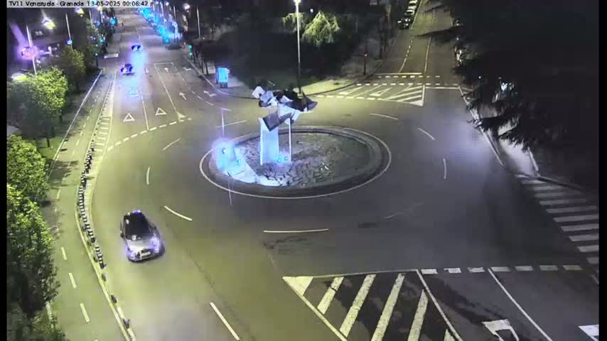 A nighttime aerial view shows a roundabout with a modern sculpture in the center, surrounded by roadways and some vehicles.