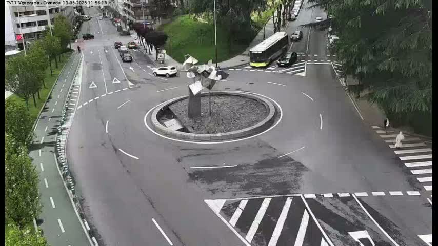 An overhead view of a roundabout with a modern sculpture in the center, several cars and a bus, and a pedestrian crossing a street.