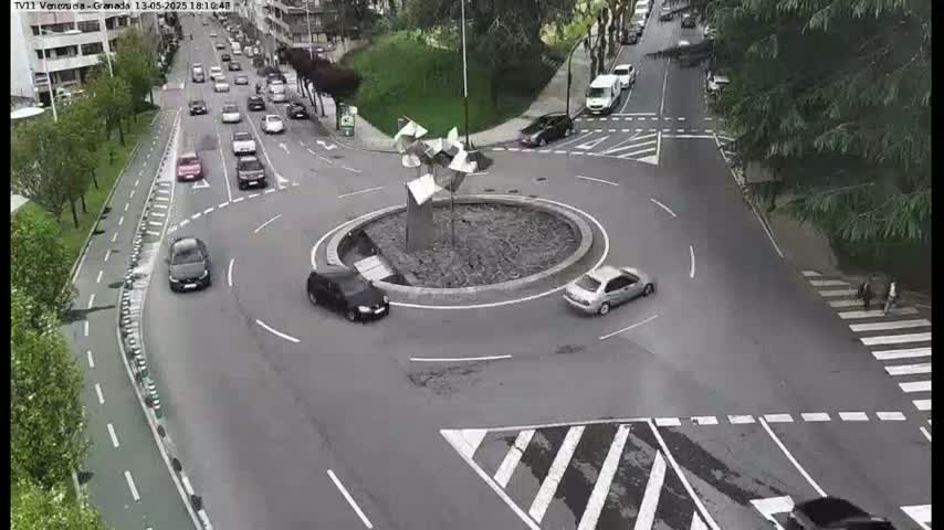 Several cars are navigating a roundabout with a modern sculpture in the center, under overcast skies.