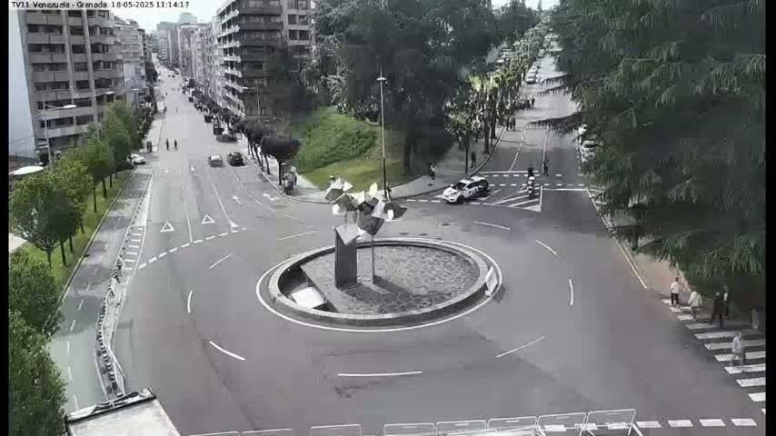 A high-angle view of a roundabout with a modern sculpture at its center, surrounded by city streets and pedestrians on a cloudy day.