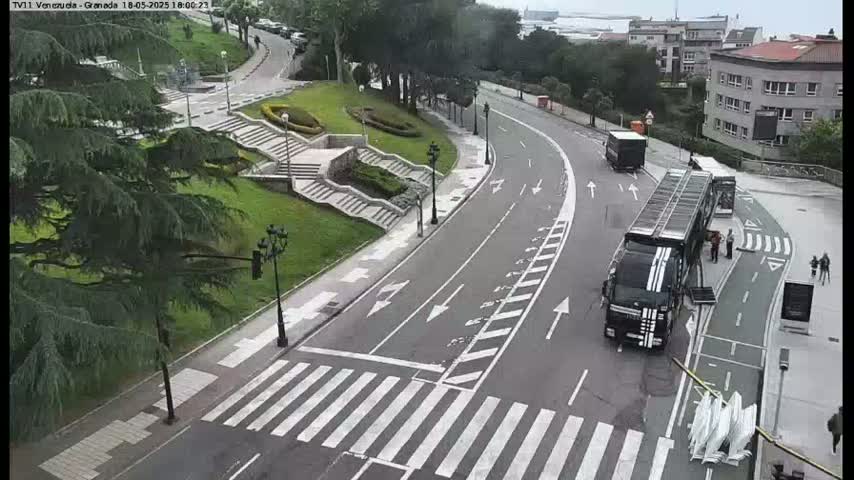 A curved road with crosswalks winds through a park-like area with stairs and landscaping, alongside a large truck and several people standing near it under a partly cloudy sky.