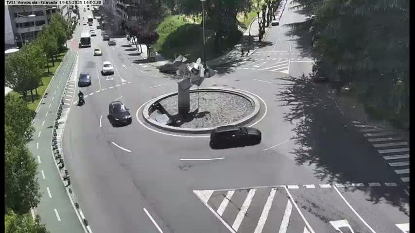 Several cars are navigating a roundabout in a city, under a partly sunny sky.