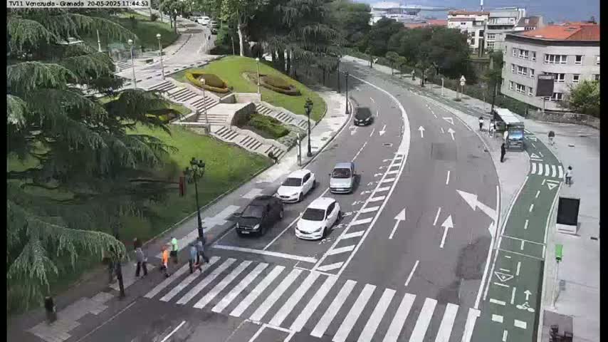 A curved road with several cars and pedestrians, next to a park with stairs and greenery, on a partly cloudy day.