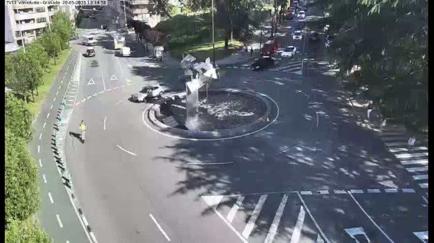A traffic circle with a modern sculpture fountain in the center is surrounded by parked and moving cars under a sunny sky.