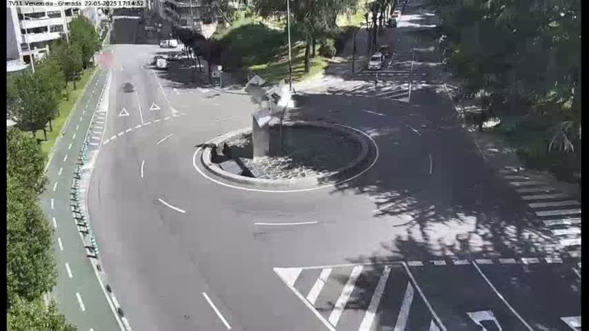 A mostly empty roundabout with a sculpture in the center is shown on a sunny day, surrounded by roads, a bike path, and trees.