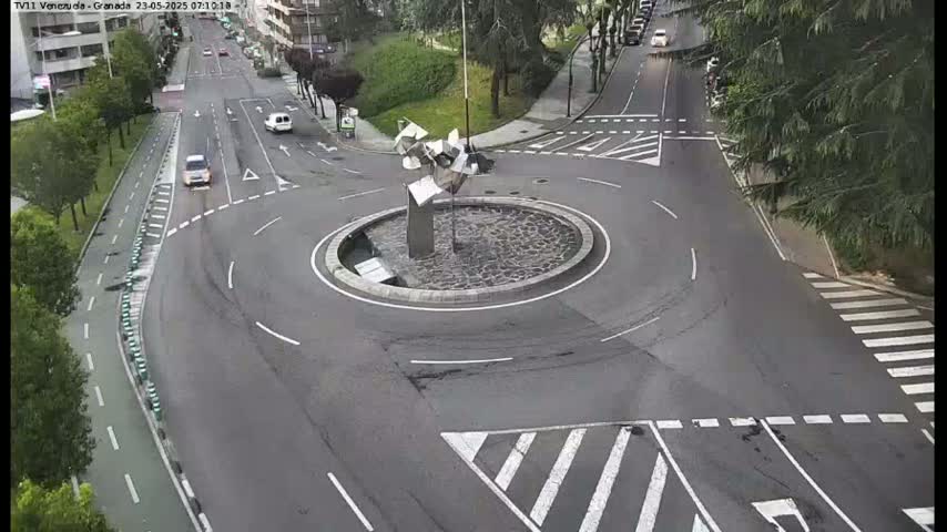 A mostly empty roundabout with a modern sculpture in the center is surrounded by roads with a few cars driving on a cloudy day.