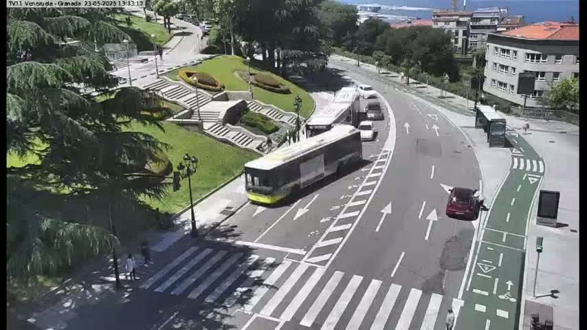 A city street scene on a sunny day shows a bus and several cars stopped at an intersection near a park with stairs and lush green landscaping.