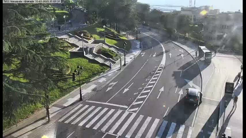 A sunny day overlooks a roadway with crosswalks, a few cars, and pedestrians near stairs leading down to a park-like area with lush green trees.