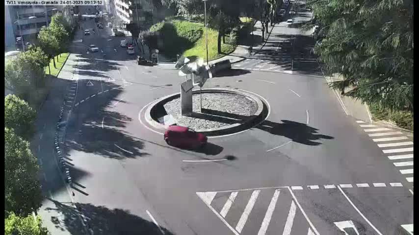 A sunny day shows a red car driving around a roundabout in a city, with some other cars visible on nearby roads.