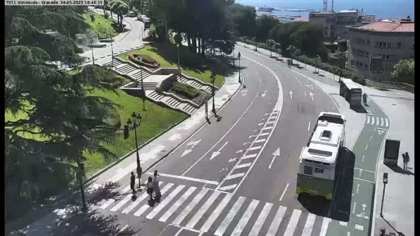 A curving road with a crosswalk, pedestrians, and two buses is seen on a sunny day, alongside a landscaped area with stairs and trees.