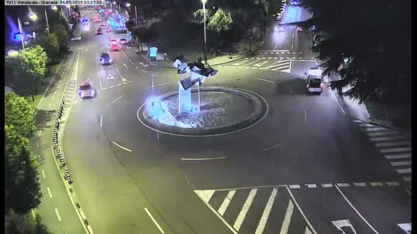 A nighttime aerial view shows a roundabout with a modern sculpture in the center, surrounded by roads with several vehicles and trees.