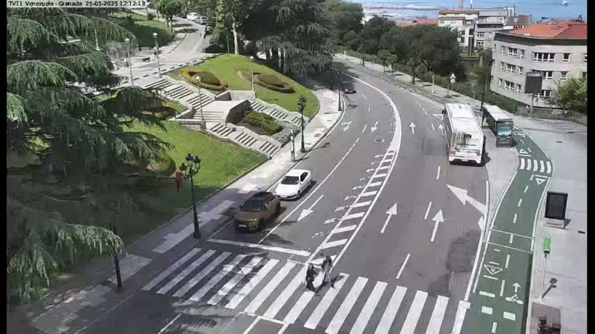 A sunny day overlooking a roadway with several cars, buses, and pedestrians crossing a crosswalk, situated near a landscaped area with stairs and greenery.