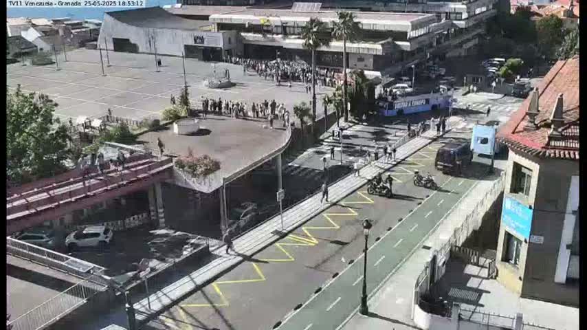 A sunny, outdoor scene shows a paved area with people walking, cars parked, and several vehicles on a road next to a pedestrian walkway that crosses a bridge over another road, with buildings in the background.