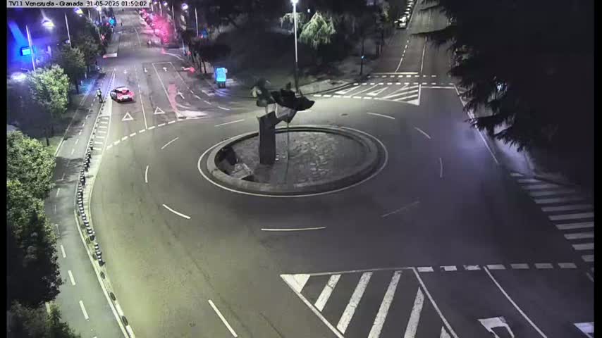 A nighttime aerial view shows a roundabout with a dark sculpture in the center, surrounded by roads and a pedestrian crossing.