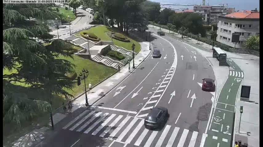 A sunny day shows a curved road with several cars and a bus, intersecting with a crosswalk near a grassy area with stairs and landscaping.