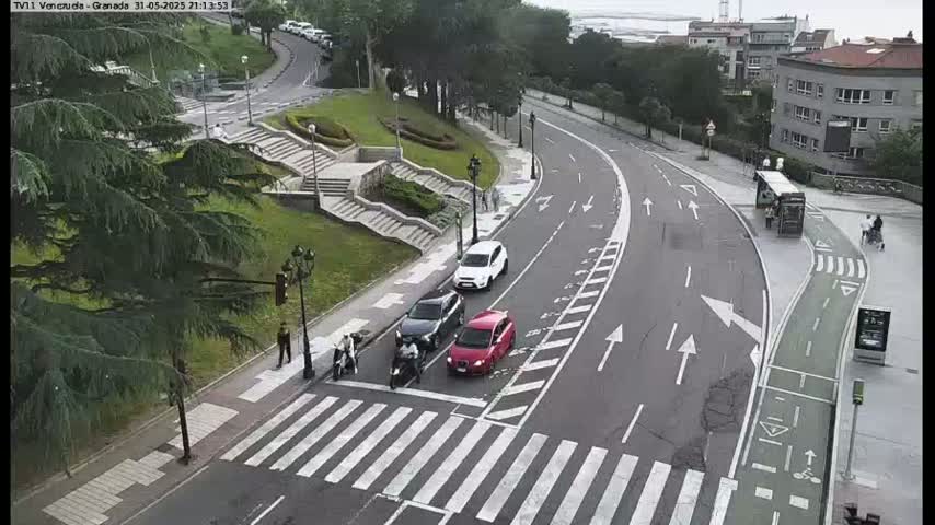 A street scene shows several cars at a crosswalk near a set of stairs, a bus stop, and some buildings, on an overcast day.
