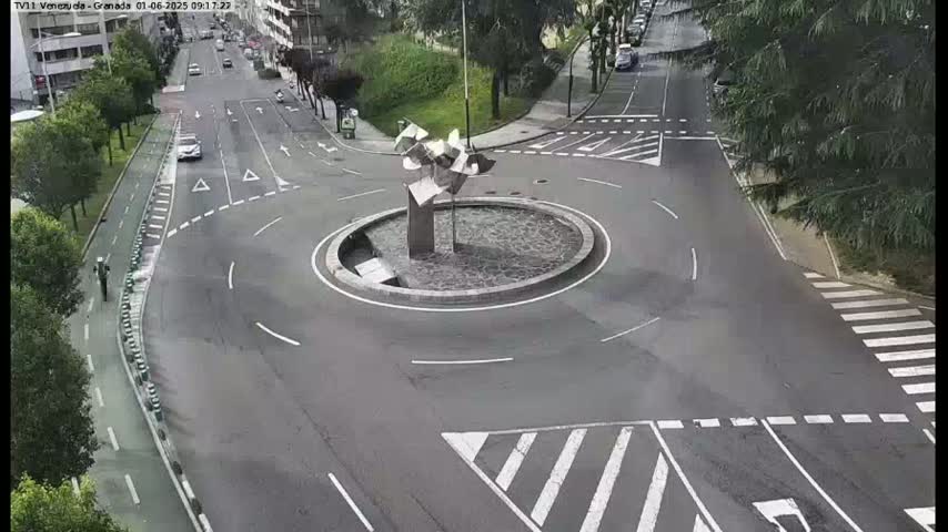 An overhead view shows a roundabout with a modern metal sculpture in the center, surrounded by roads with light traffic under overcast skies.