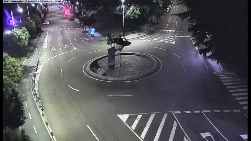 A nighttime aerial view shows a roundabout with a dark sculpture in the center, surrounded by roadways with minimal traffic.