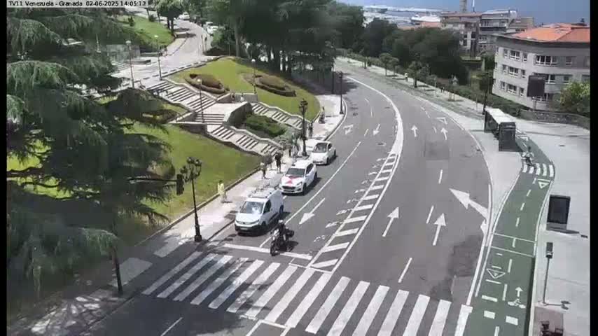 A sunny day shows a curved road with several cars and a motorcycle near a pedestrian crossing and stairway leading to a grassy area with buildings in the background.