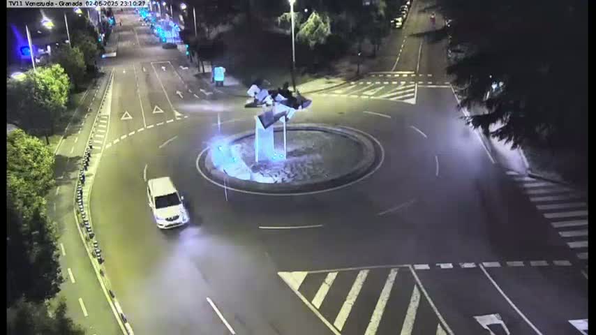A white car drives through a roundabout at night, illuminated by streetlights.
