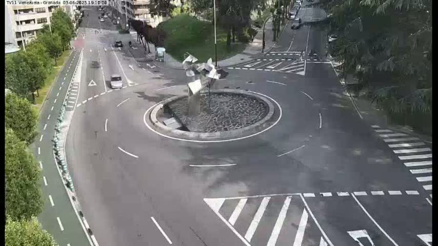 A roundabout with a modern sculpture in the center is surrounded by roads and a bike lane, with a few cars visible on a partly sunny day.