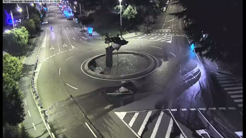 A dark, wet roundabout at night with a sculpture in the center, surrounded by roads with light traffic.