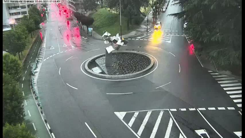 A wet roundabout with a modern sculpture in the center is surrounded by roads with sparse traffic under a cloudy sky.