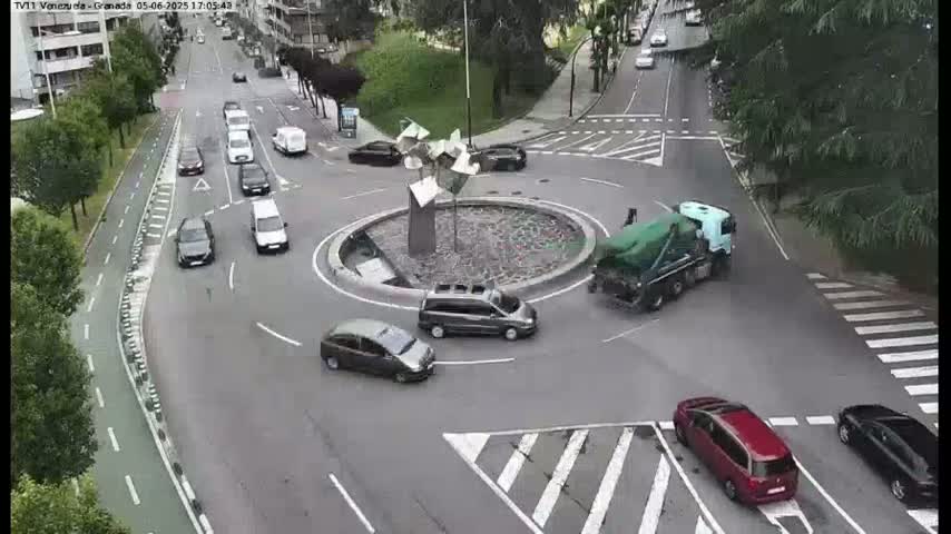 Several cars are navigating a roundabout in a city, with a cement mixer truck exiting it, under partly sunny conditions.