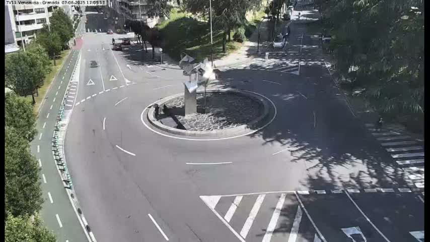 A roundabout with a modern sculpture in the center is surrounded by roads and trees under sunny conditions.