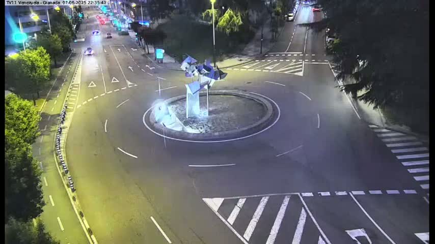 A nighttime aerial view shows a roundabout with a modern sculpture in the center, surrounded by roads with minimal traffic.