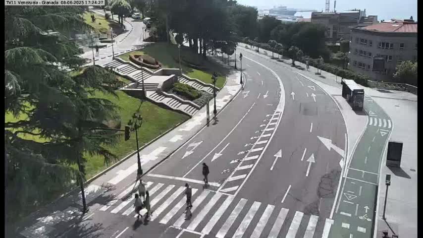 A mostly empty road curves through a park-like setting on a sunny day, with pedestrians crossing a crosswalk and a bus parked at a bus stop.