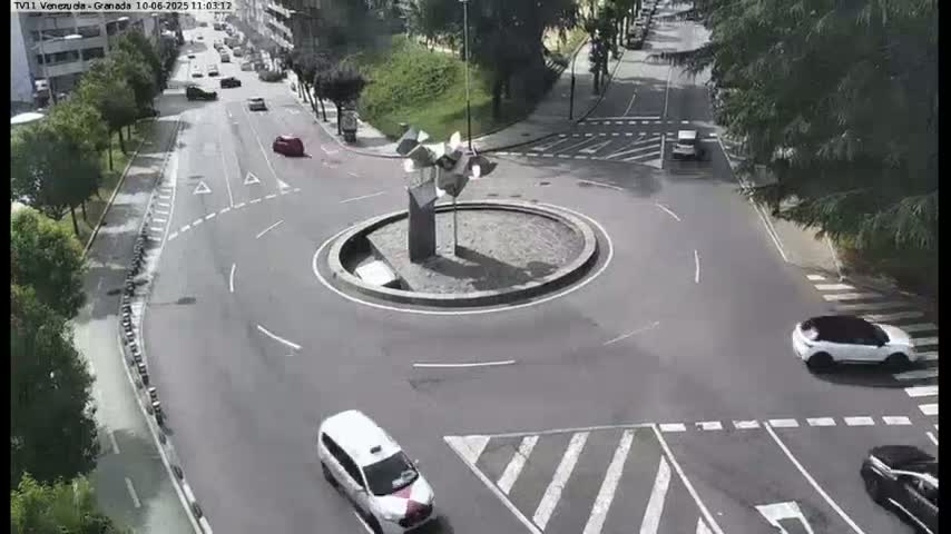 A high-angle view shows a traffic circle with several cars moving around it on a sunny day, and a modern sculpture in the center.