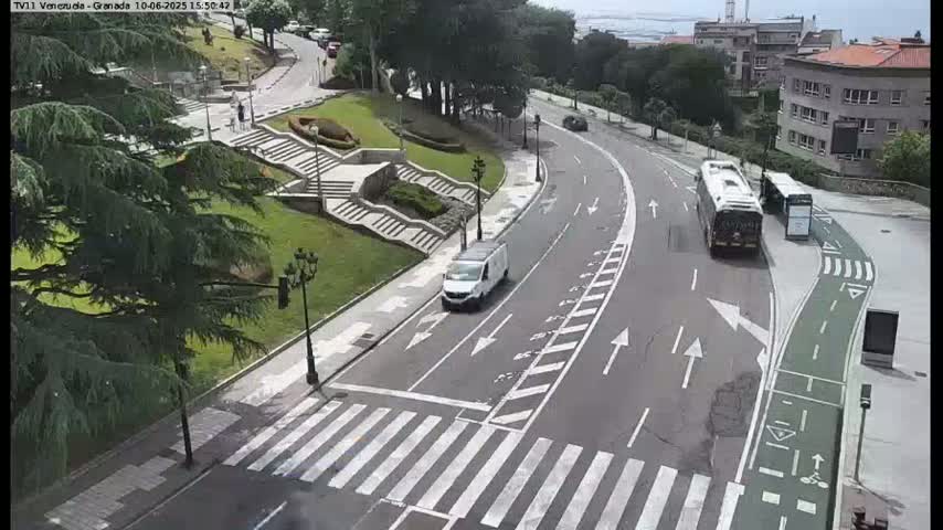 A curved road with a pedestrian crossing and a bus stop, surrounded by green landscaping and buildings, on a sunny day.