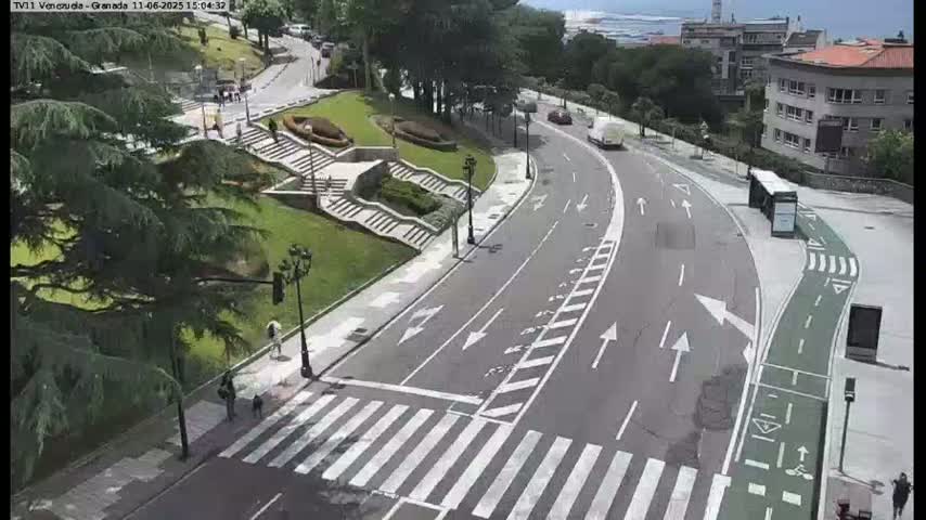 A curved road with pedestrian crossings and bike lanes winds through a green, landscaped area with stairs and buildings visible in the background on a sunny day.