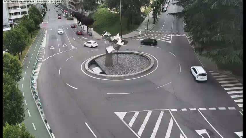 A traffic circle with several cars driving around it and a modern sculpture in the center, under a partly cloudy sky.