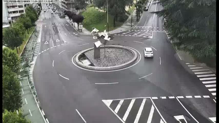 A wet roundabout with a modern sculpture in the center, surrounded by roads with a few cars visible.