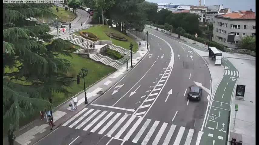 A curved road with a crosswalk and bike lane winds through a green space, with pedestrians and a single car visible on a partly cloudy day.