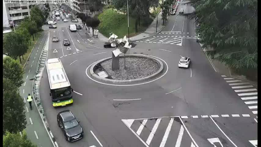 A roundabout with a modern sculpture in the center, surrounded by cars and a bus, on an overcast day.
