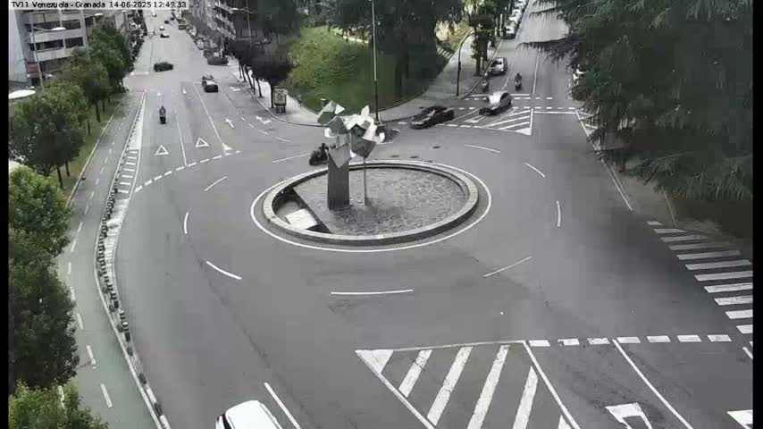 A roundabout with a modern sculpture in the center is surrounded by roads with several cars and a motorcycle, under a partly cloudy sky.