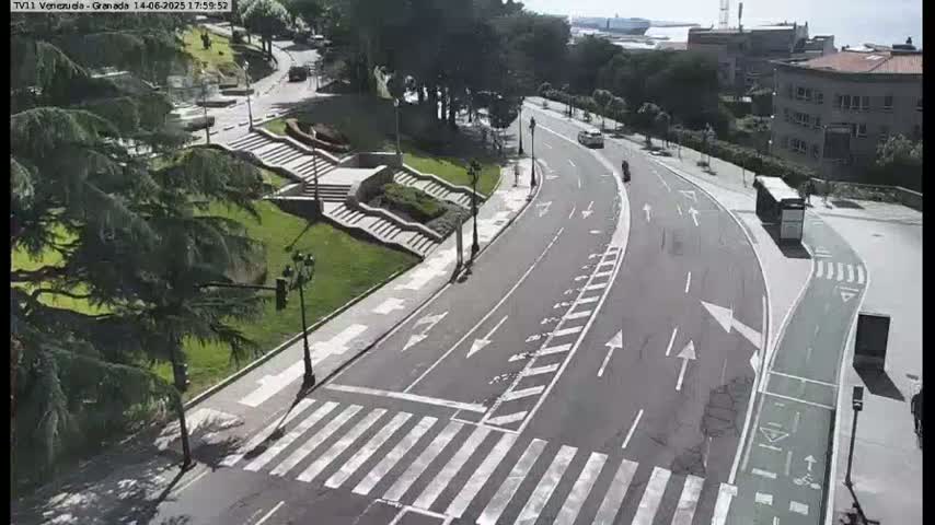 A mostly empty road curves through a park-like area on a sunny day, with a pedestrian crossing and a bike lane visible.