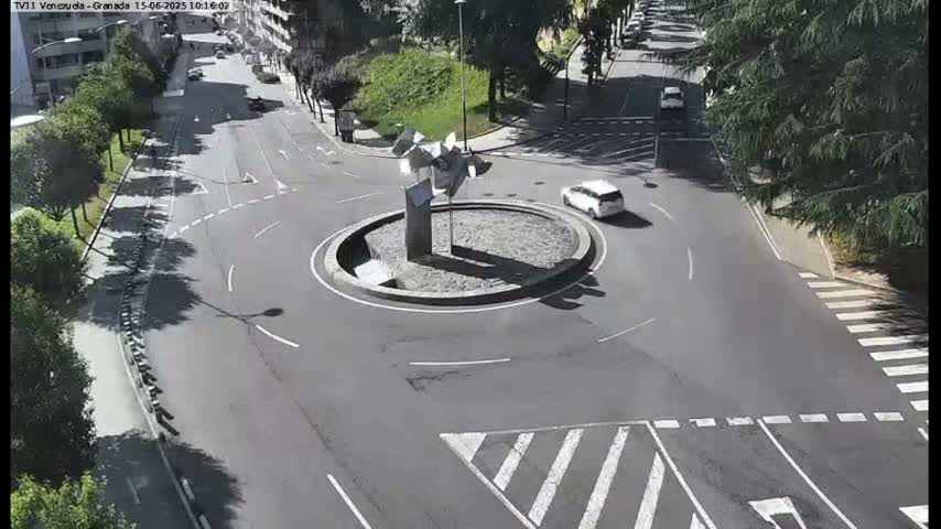 A sunny day shows a traffic roundabout with a modern sculpture in the center, a few cars, and surrounding roads with crosswalks.