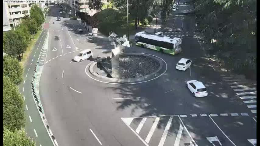 A sunny day shows a roundabout with several cars and a bus, surrounded by trees and buildings.