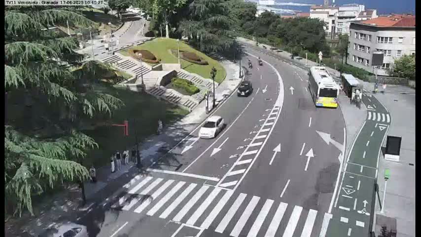A curving road with a pedestrian crossing and a bus stop is seen on a sunny day, with a few cars and pedestrians visible.