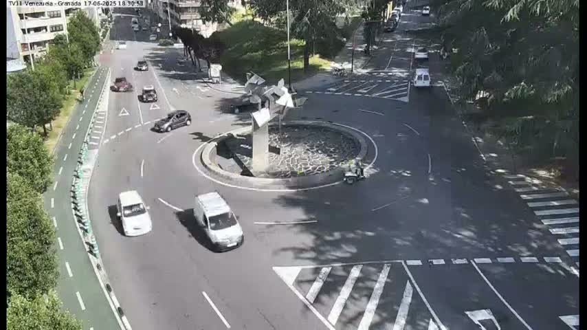 Several cars navigate a roundabout in a sunny urban setting, with a sculpture at its center and surrounding greenery.