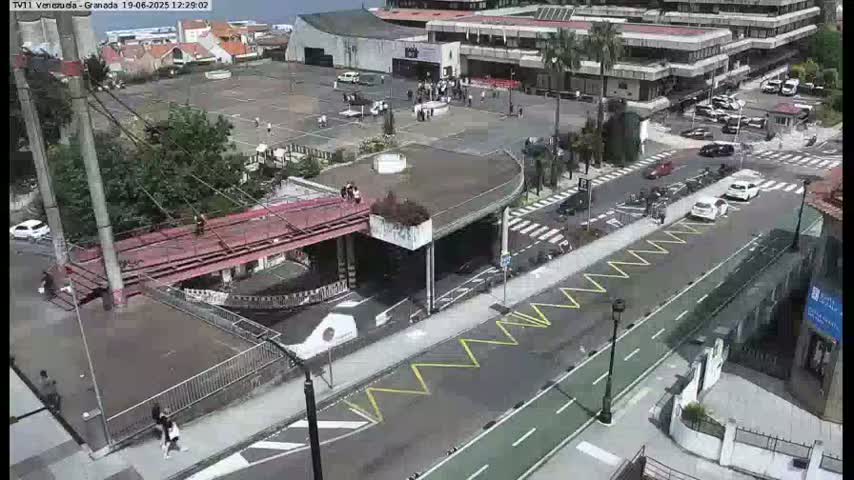 An aerial view shows a plaza with a red pedestrian bridge, a roadway with parked cars and a bike lane, and several buildings on a sunny day.