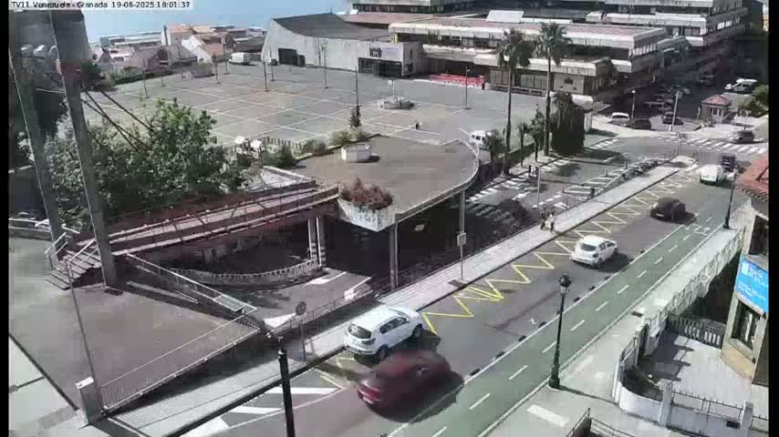 A mostly empty parking lot and roadway are visible from an elevated viewpoint on a sunny day, with several cars driving on the street and a pedestrian bridge present.
