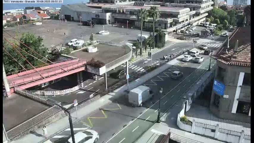 A high-angle view of a city street scene on a sunny day, showing a parking lot, a bridge, buildings, and several cars.