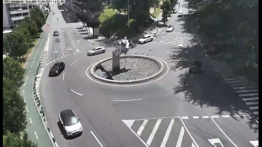 Several cars are driving around a roundabout with a stone fountain in the center, on a sunny day.