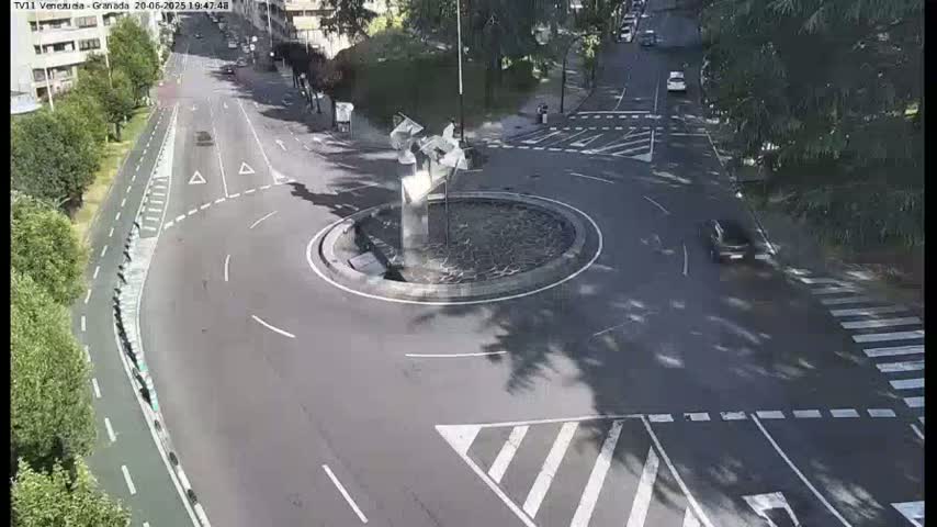 A roundabout with a modern sculpture in the center is surrounded by a paved road with crosswalks and bike lanes, under a sunny sky.