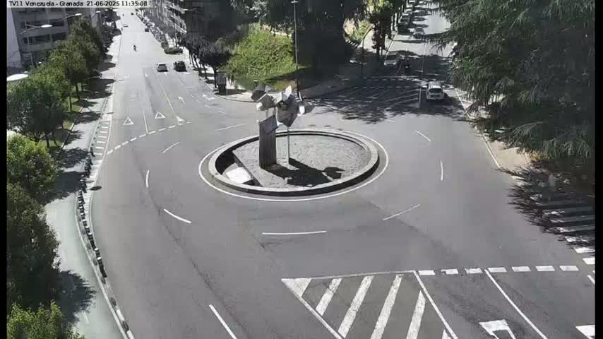 A sunny overhead view shows a circular traffic island with a sculpture in the center of a roadway intersection, surrounded by several cars and trees.